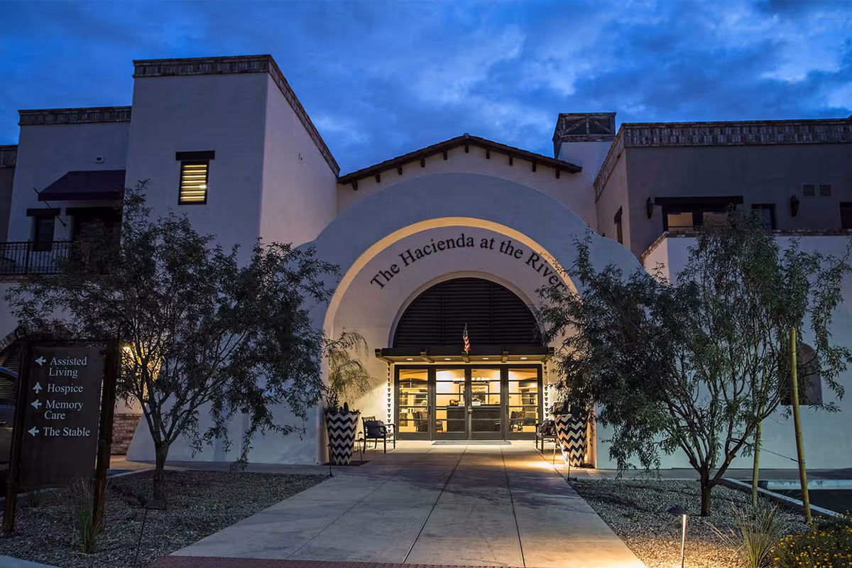 Exterior front entrance of The Hacienda at the River facility at dusk, featuring an arched doorway with glass doors, illuminated from inside. There are two large decorative planters on either side of the entrance and trees flanking the walkway leading to the door. A sign on the left indicates directions to Assisted Living, Hospice, Memory Care, and The Stable.