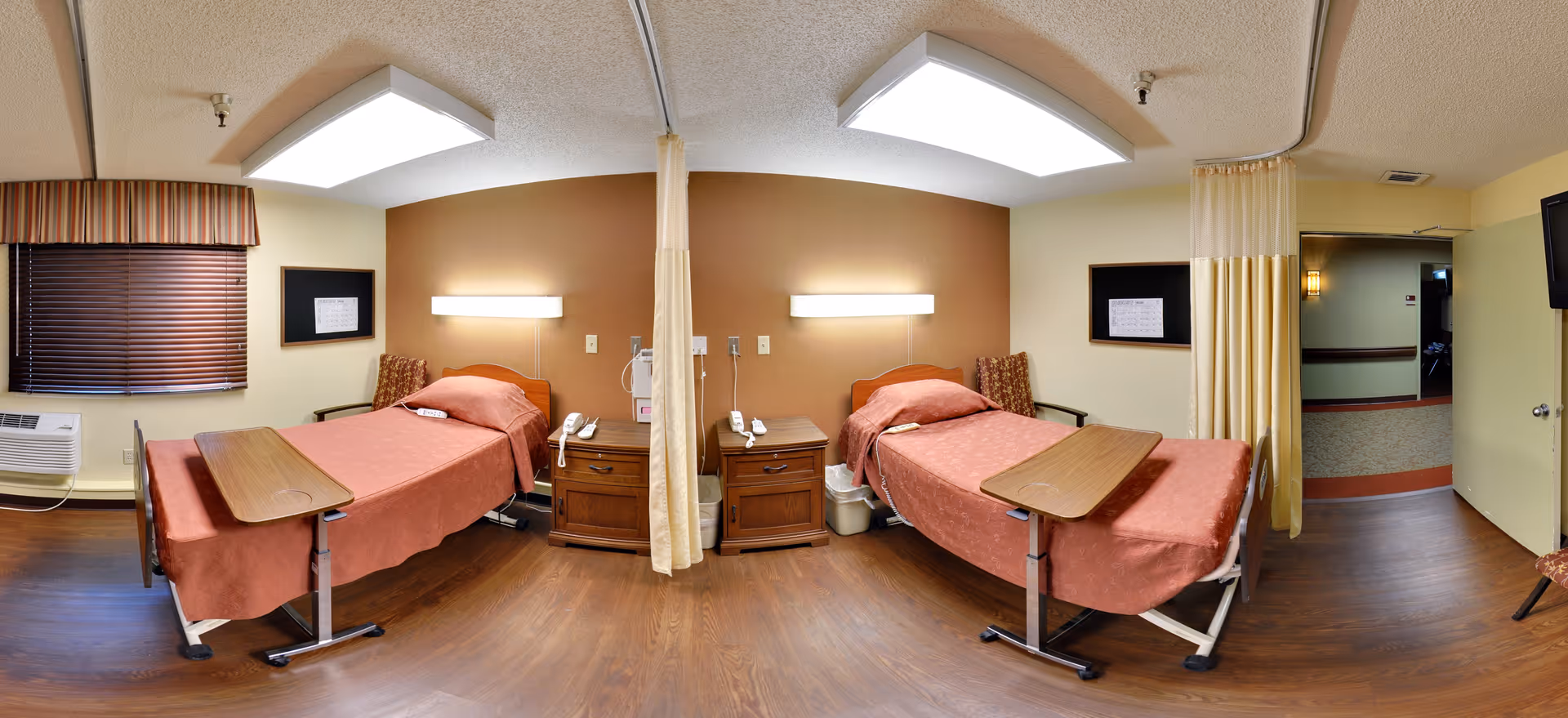 A shared hospital or senior care facility room with two beds separated by a privacy curtain. Each bed has a wooden overbed table, a bedside table with a telephone, and a wall-mounted light above the headboard. The room has wood flooring, a window with blinds, and a TV mounted on the wall near the door.