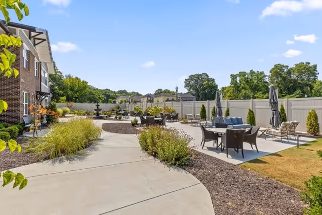 Sunny outdoor courtyard with paved walkways, patio seating and umbrellas, landscaping, and a brick building at the side.