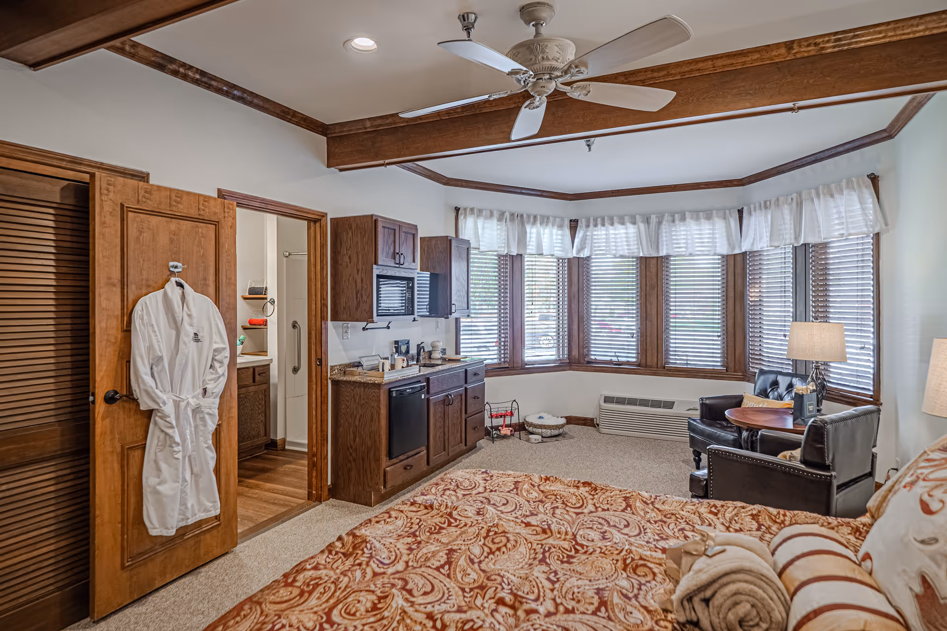 A cozy retirement community room featuring a bed with patterned bedding in the foreground, a small kitchenette with wooden cabinets and appliances, a sitting area with two leather chairs and a small round table near a large bay window with wooden blinds and white valances, a ceiling fan with light, and a bathroom visible through an open door with a white robe hanging on the door.