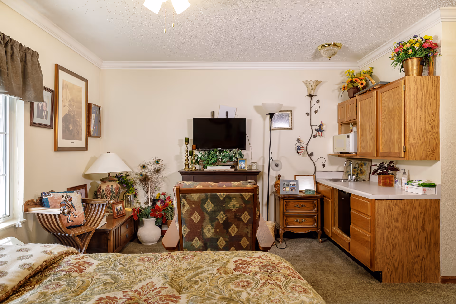A cozy room featuring a bed with floral bedding in the foreground, a patterned armchair facing a wall-mounted TV, a small kitchenette with wooden cabinets, a microwave, and a sink. The room is decorated with framed pictures, a table lamp, various plants, and flowers. There is a window with a brown valance on the left side.