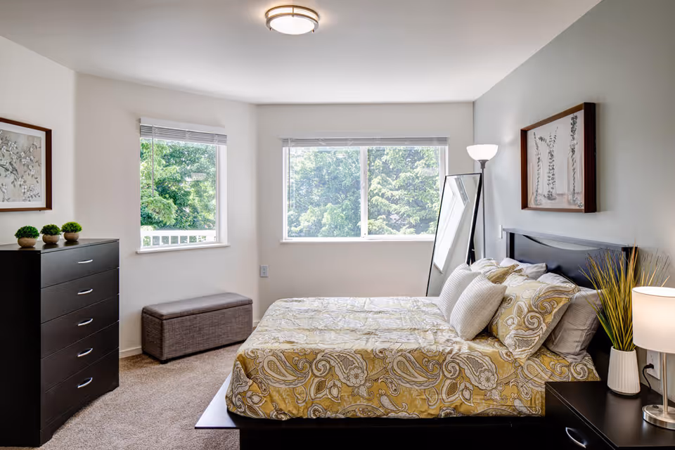 A bright bedroom with a large window showing green trees outside. The room features a bed with a yellow and white patterned comforter and multiple pillows. There is a black dresser with three small potted plants on top, a gray storage bench under a smaller window, a floor lamp, a standing mirror, a nightstand with a lamp and a plant, and framed artwork on the walls.