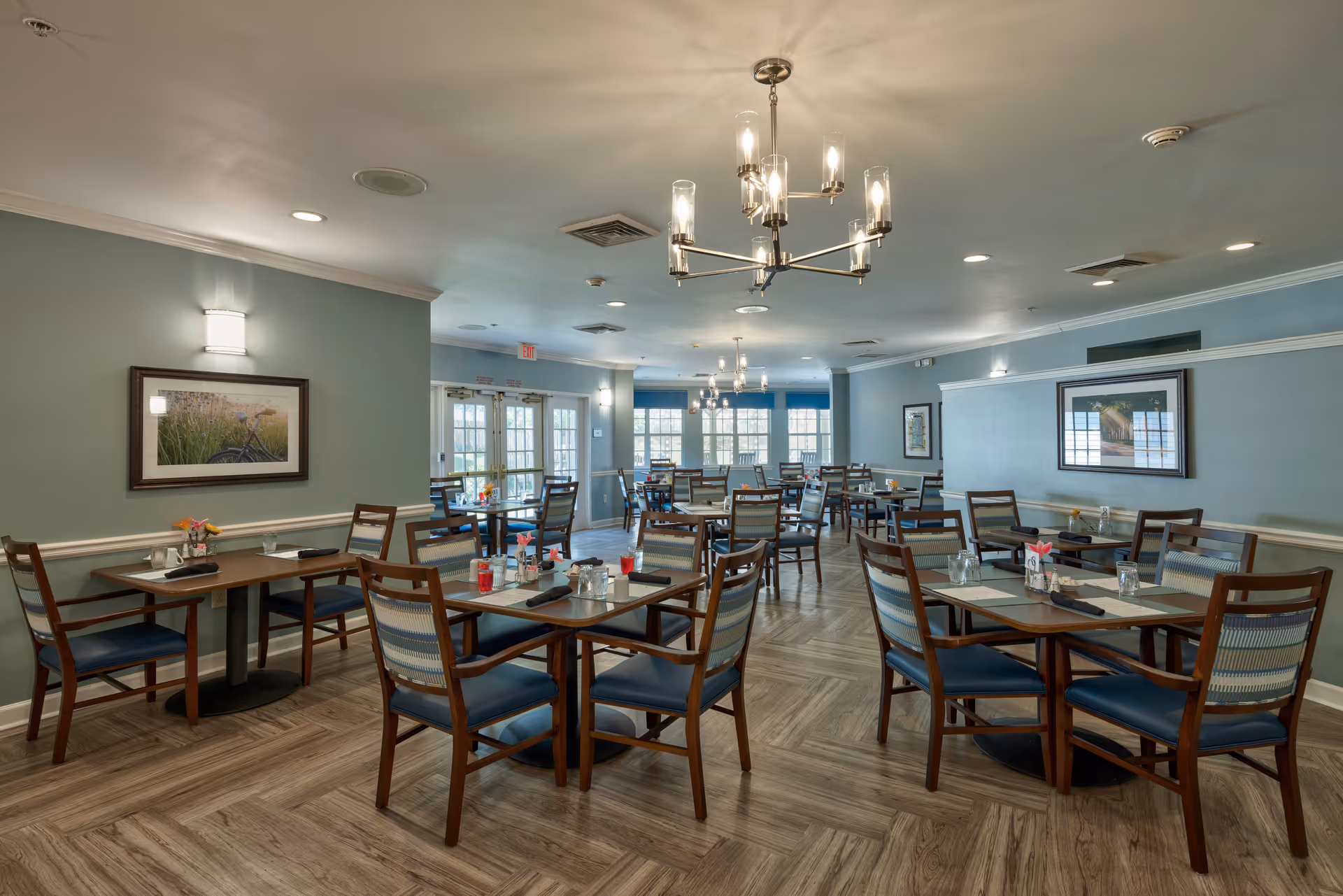 A spacious dining room with multiple wooden tables and chairs arranged neatly. Each table is set with placemats, napkins, glasses, and small flower vases. The room has light blue walls adorned with framed pictures, wood-patterned flooring, and modern ceiling light fixtures. Large windows and glass doors at the far end allow natural light to enter the room.