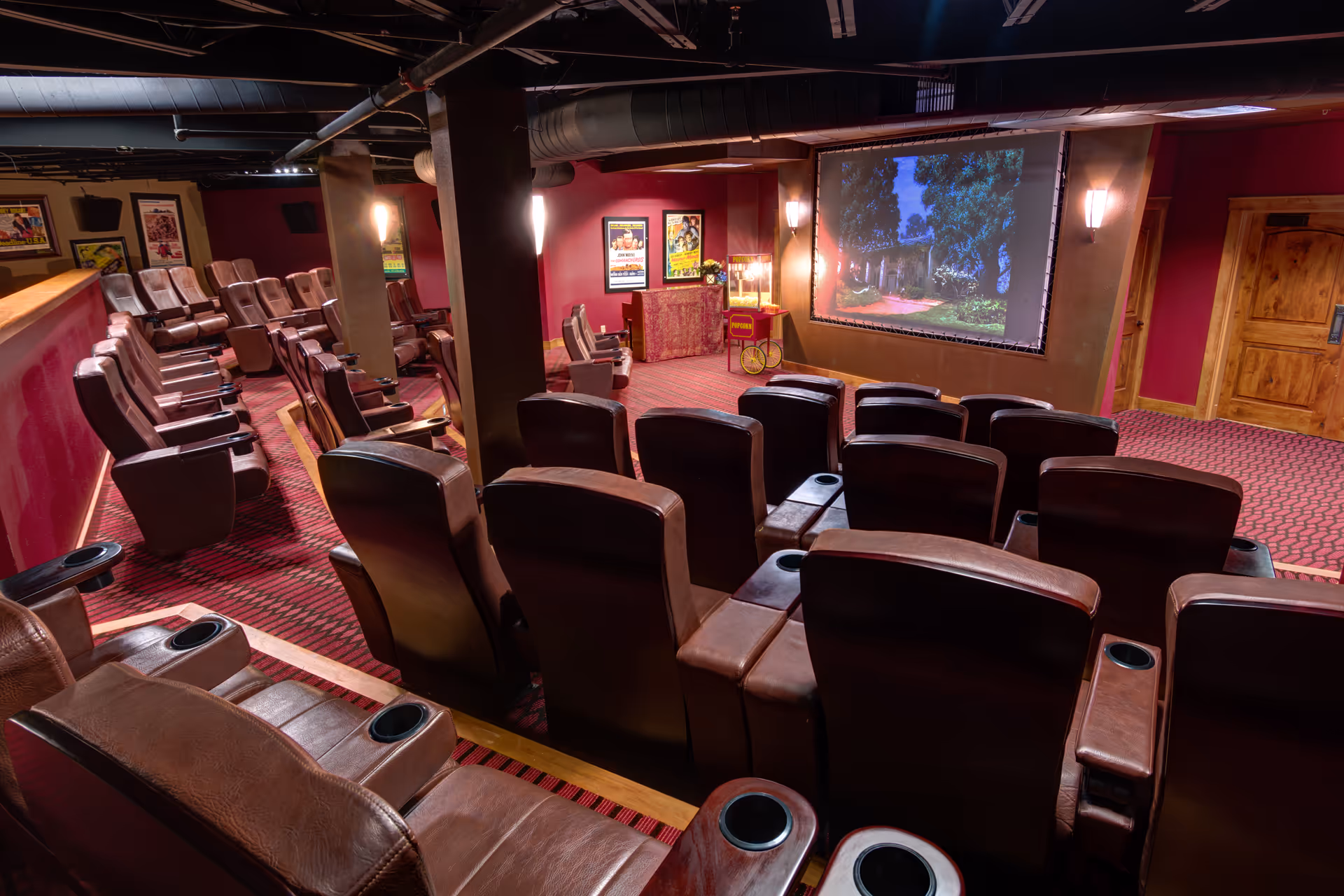 Interior view of a small movie theater room with multiple rows of brown leather recliner seats facing a large screen showing an outdoor scene. The room has red walls, carpeted floor, wooden doors, and movie posters on the walls. There is a popcorn machine near the screen.