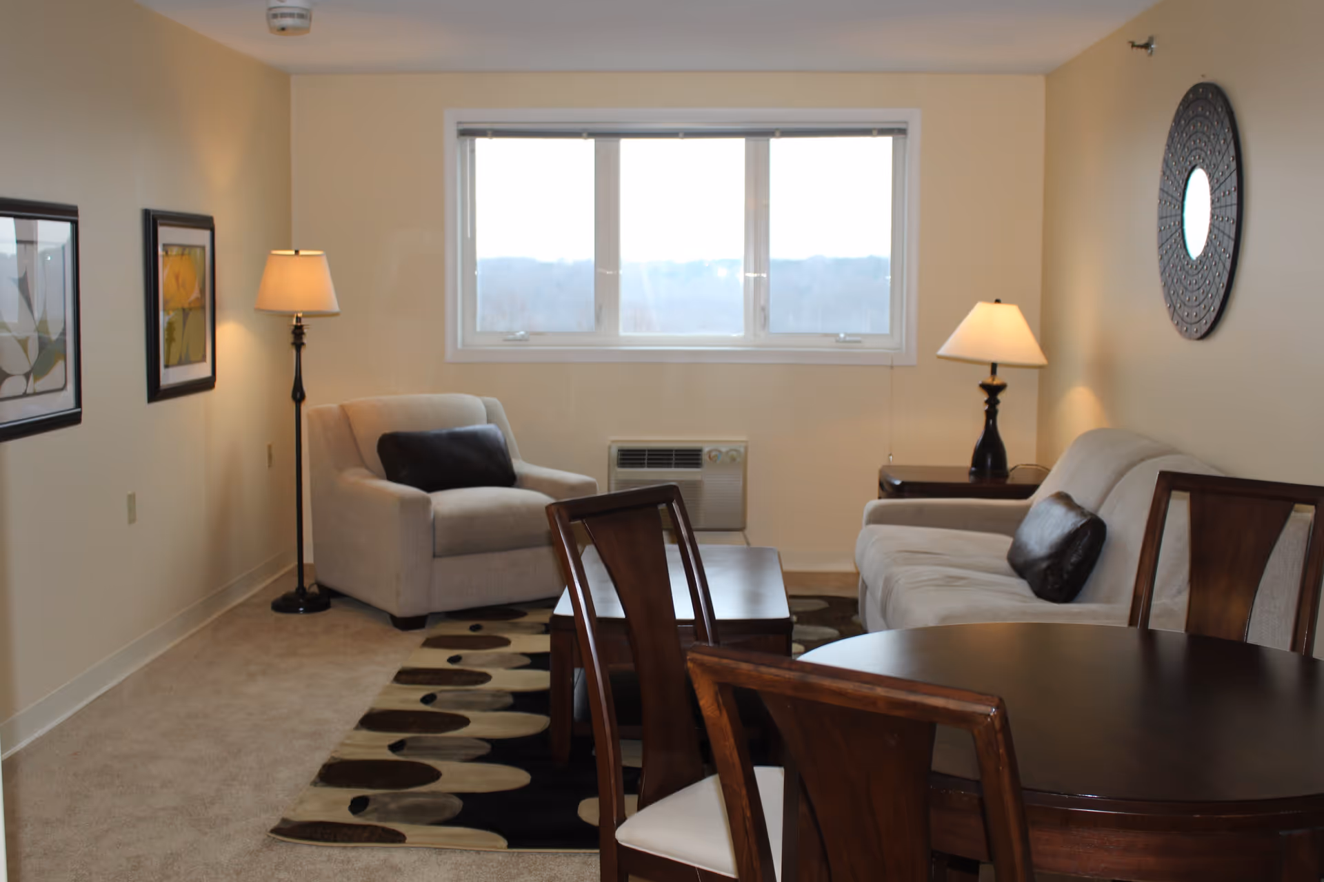 A cozy living room with a beige armchair and sofa, both with dark cushions. There are two lamps, one floor lamp next to the armchair and one table lamp on a side table next to the sofa. A round wooden dining table with four chairs is in the foreground. The room has a window with a view of the outdoors, a patterned rug on the carpeted floor, two framed pictures on the left wall, and a decorative round mirror on the right wall.