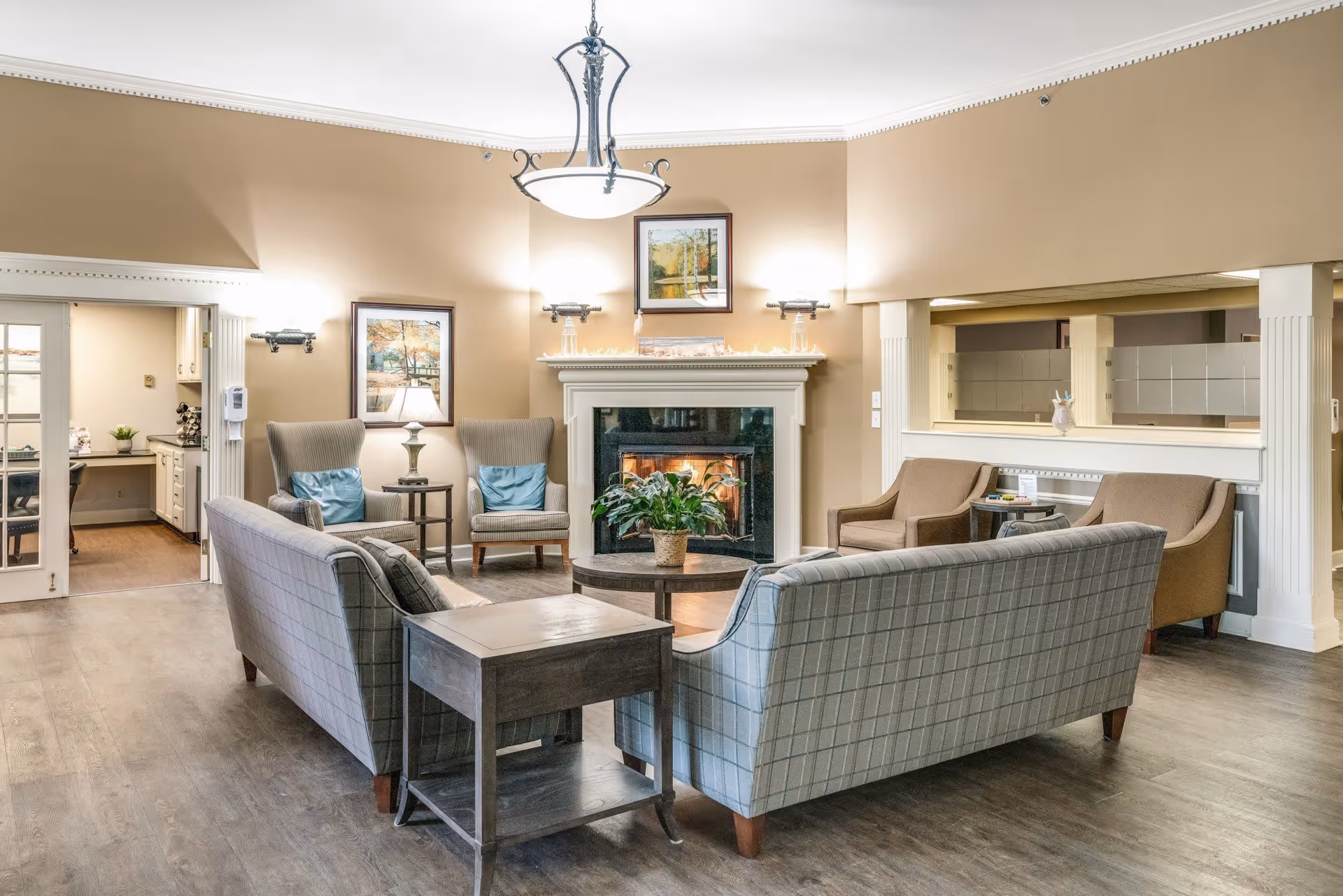 A cozy living room area in a senior living facility named Shorehaven, featuring two plaid sofas facing each other with a wooden side table between them, two armchairs with blue cushions near a lit fireplace, a round coffee table with a potted plant, and warm beige walls adorned with framed artwork. The room has wood flooring and a chandelier hanging from the ceiling. A kitchen area is visible through an open doorway on the left.