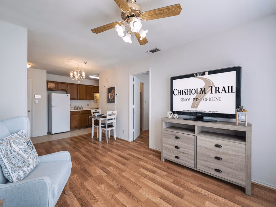 Interior view of a senior living apartment featuring a light blue armchair with a patterned cushion, a wooden floor, a small dining table with two white chairs, a kitchen area with wooden cabinets and a white refrigerator, and a TV on a wooden stand displaying the Chisholm Trail Senior Living of Keene logo.