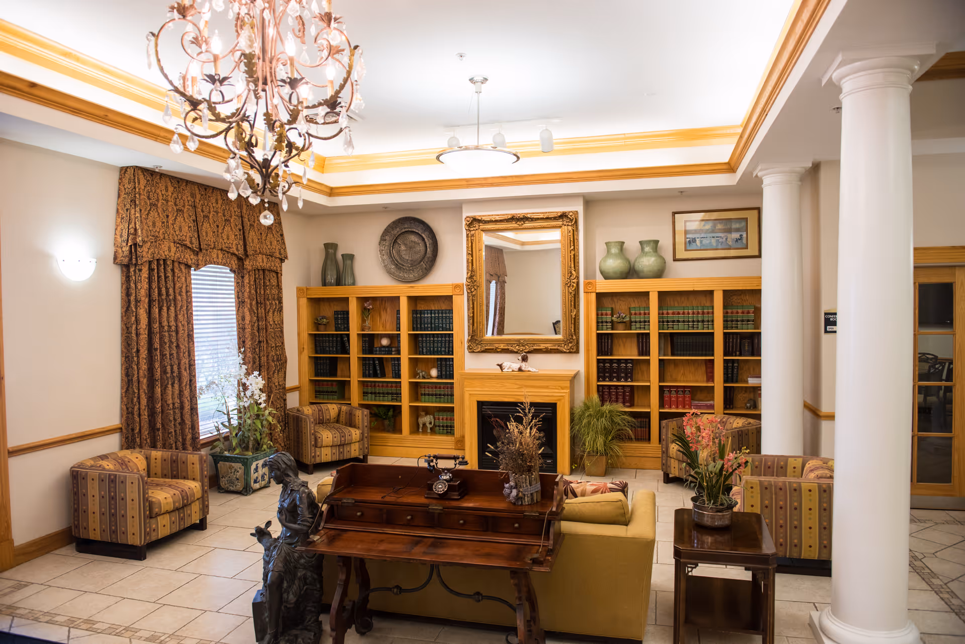 A warmly decorated lounge with a chandelier, seating, bookshelves and a central coffee table in front of a fireplace and mirror.