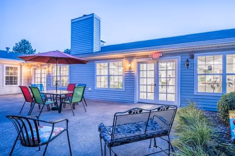 Outdoor patio area at The Courtyard at Fremont with metal chairs and tables, one table shaded by a red umbrella, surrounded by a building with multiple windows and a door with an exit sign above it, some greenery on the side.