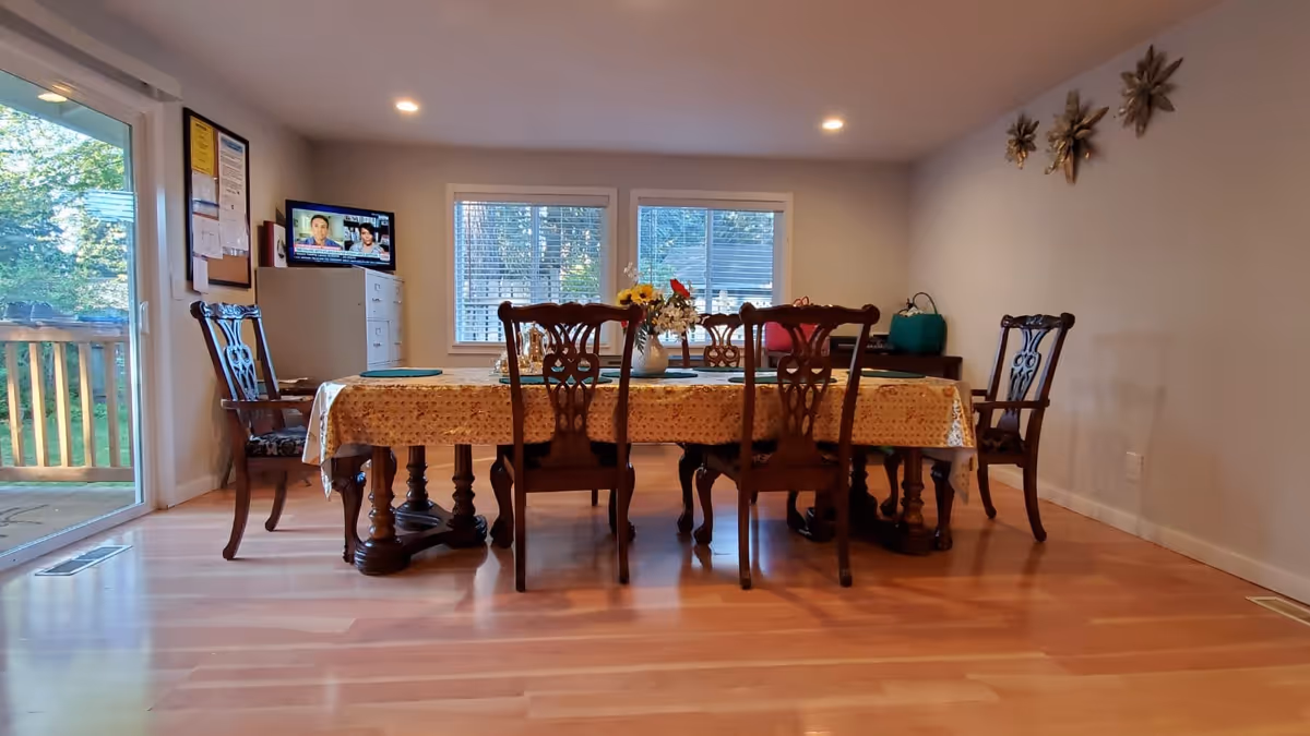 Bright dining room with a long wooden table covered by a tablecloth, six ornate chairs, windows and a sliding glass door to a deck.