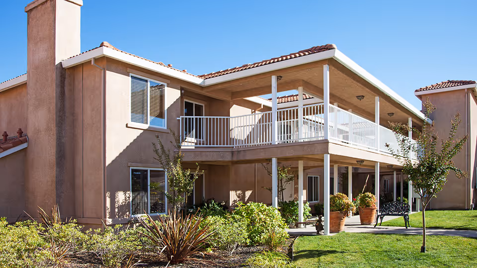 Exterior view of a two-story residential building with beige stucco walls and a red tile roof. The building features a covered balcony with white railings on the upper floor and a covered patio area below. There are several windows, small trees, shrubs, and a well-maintained lawn in the foreground under a clear blue sky.