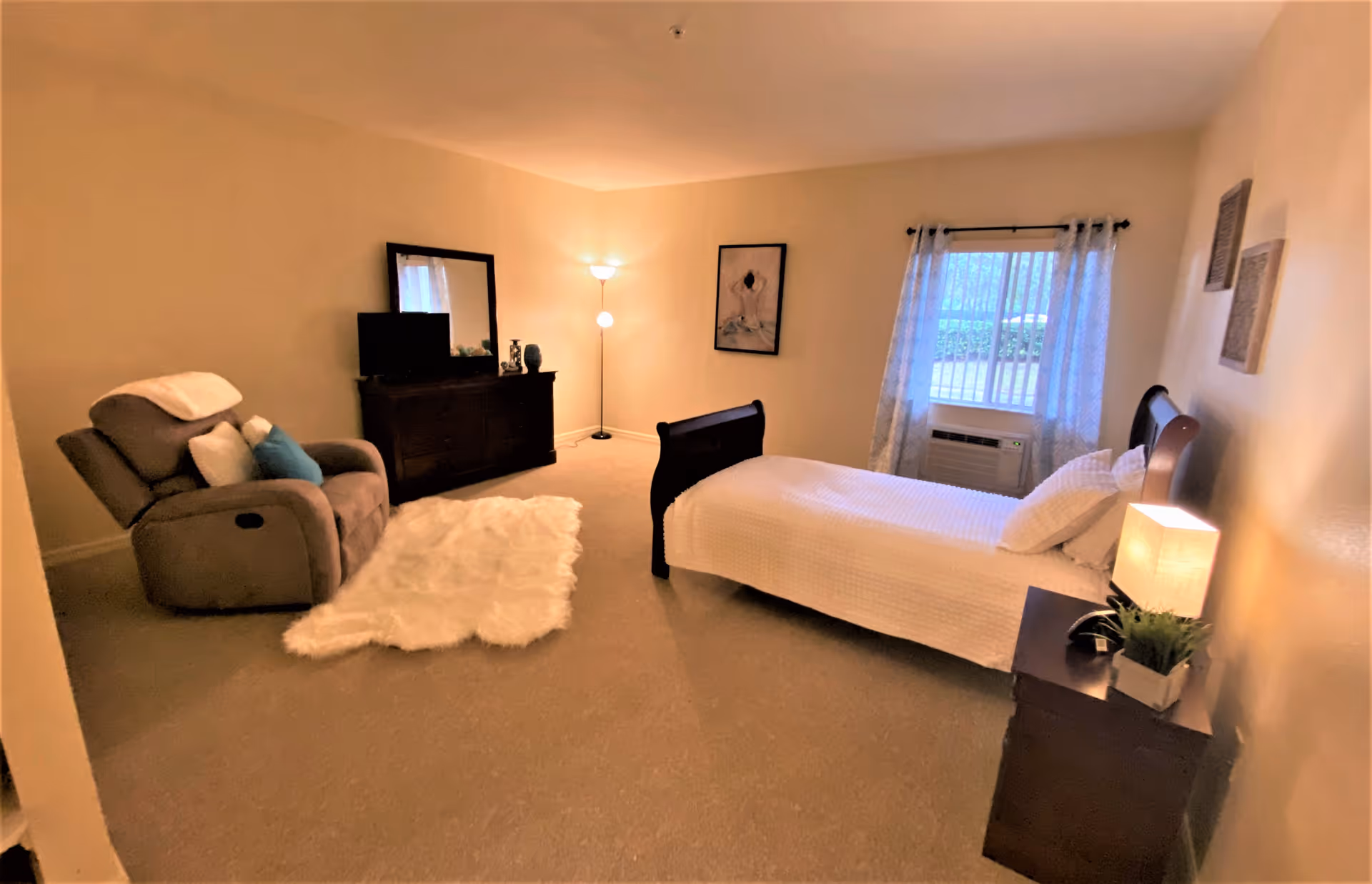 A cozy bedroom with a single bed covered in white bedding, a wooden nightstand with a lamp and a small plant, a window with sheer curtains, a dark wooden dresser with a mirror and TV on top, a floor lamp in the corner, a comfortable recliner chair with pillows, and a white fluffy rug on the carpeted floor.