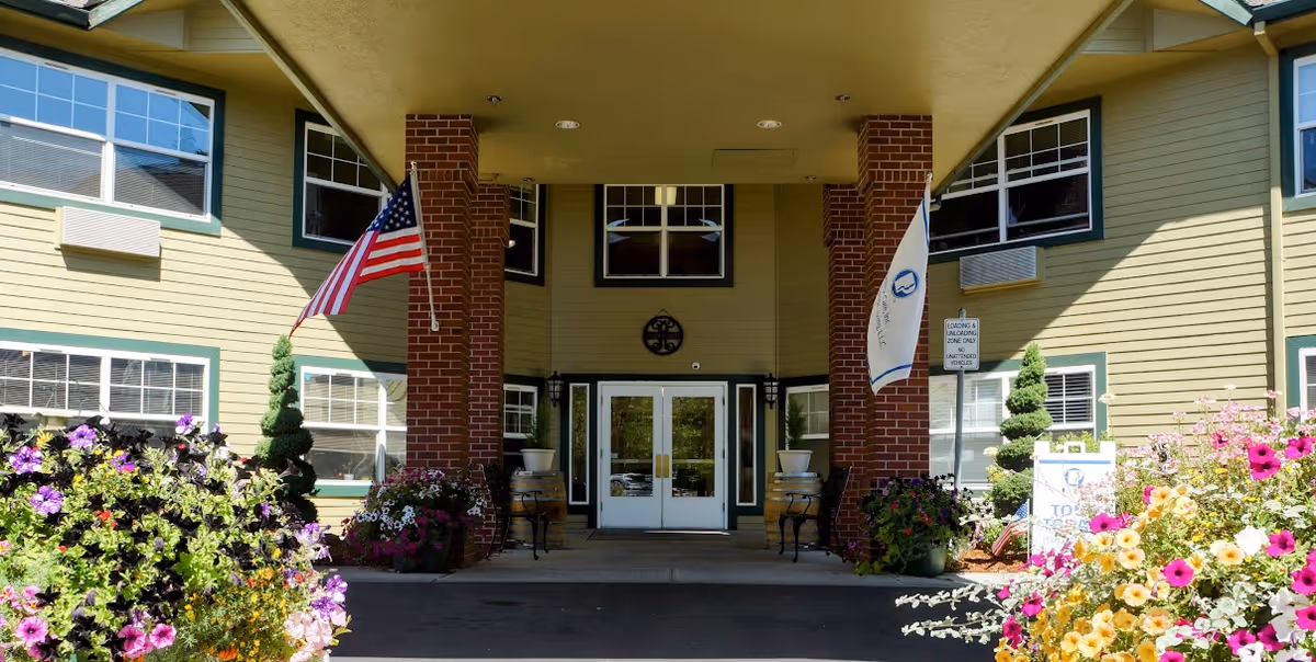 Entrance of Prestige Senior Living Riverwood featuring a covered driveway supported by brick columns, two flags on either side, and colorful flower beds in the foreground.