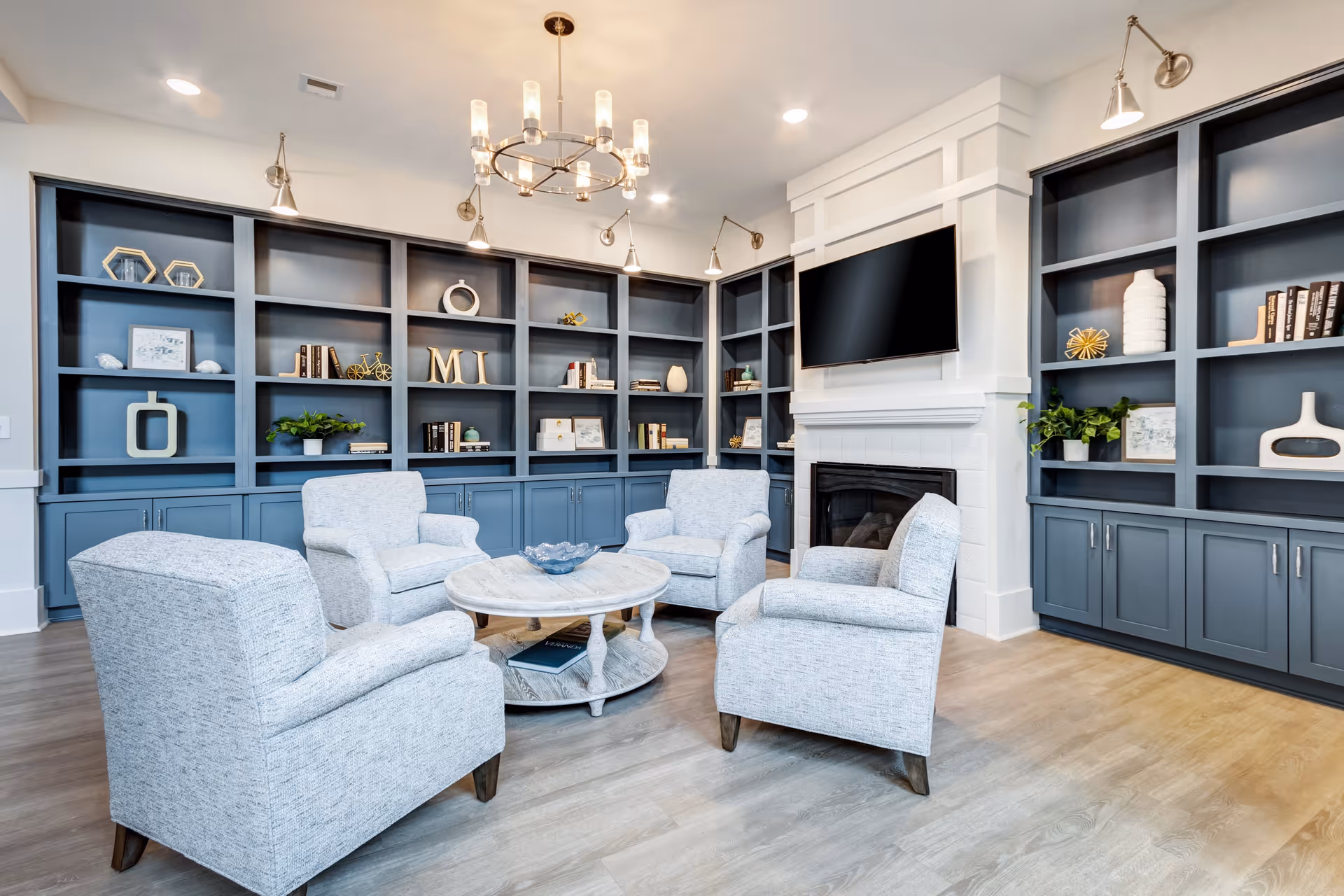 A cozy living room with four light gray upholstered armchairs arranged around a round wooden coffee table. Behind the chairs is a wall of built-in blue shelves decorated with books, plants, and decorative items. A flat-screen TV is mounted above a white fireplace. The room has light wood flooring and modern lighting fixtures on the ceiling and walls.