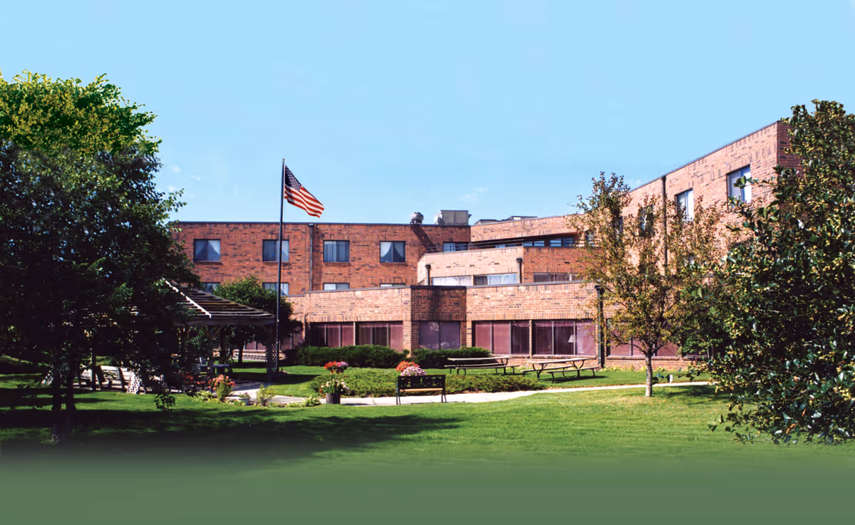 Exterior view of a brick senior living facility building with multiple windows, surrounded by green grass, trees, and a pathway. There is an American flag on a flagpole and several benches and picnic tables in the garden area.