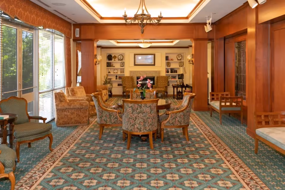 A spacious, well-lit living room with patterned carpet, floral upholstered chairs around a wooden table, armchairs, and large windows with blinds letting in natural light. The room features wooden paneling, built-in shelves with decorative items, and a chandelier hanging from the ceiling.
