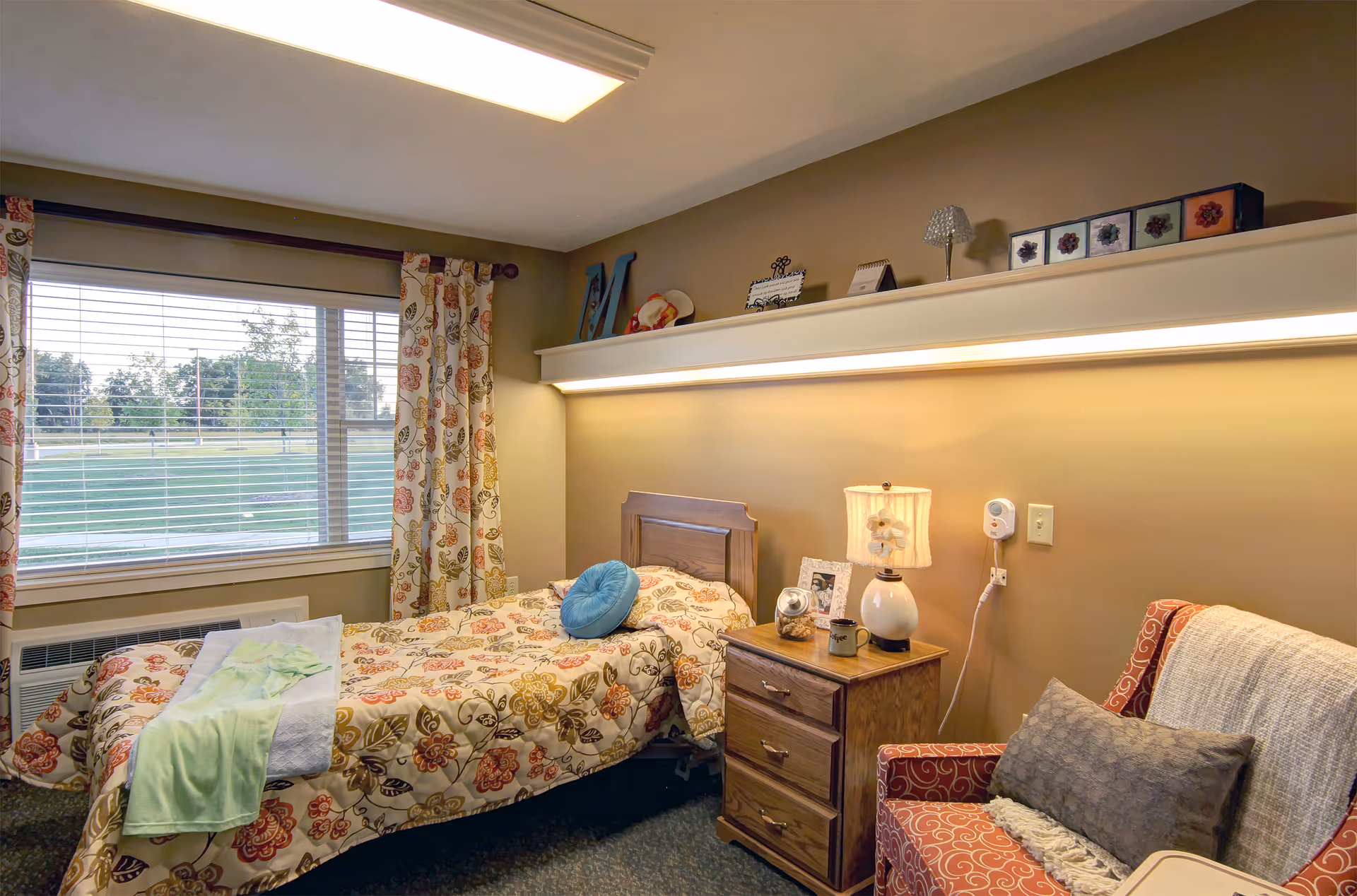A cozy senior living bedroom with a single floral bed, nightstand and lamp, armchair, and a large window overlooking a lawn.