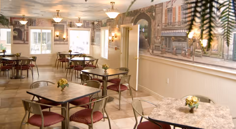 A dining room with multiple square tables, each surrounded by four chairs with red cushions. Small flower arrangements are placed on each table. The walls feature a mural depicting a quaint street scene with buildings and storefronts. The room is well-lit with ceiling lights and wall sconces, and windows with white blinds allow natural light to enter.