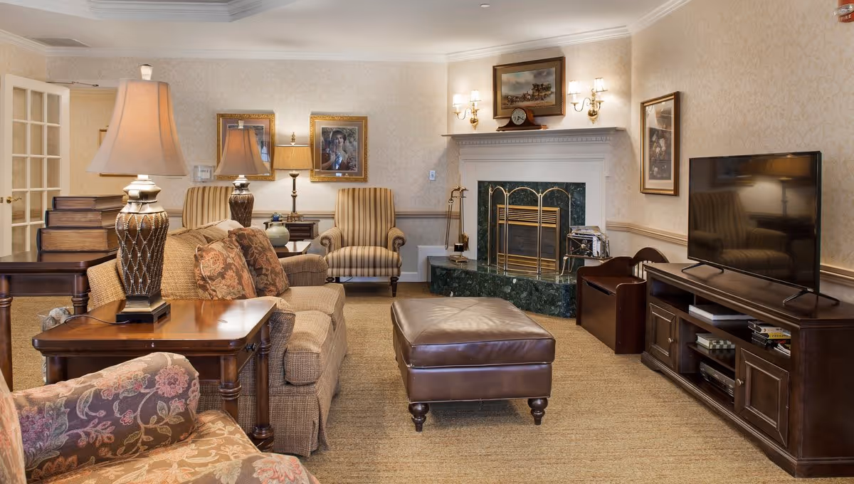 Cozy, traditionally furnished living room with upholstered sofas and armchairs, a leather ottoman, fireplace, lamps, and a TV.