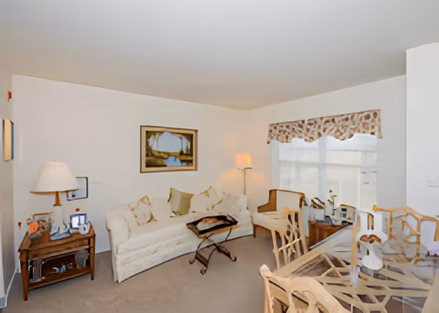 A cozy living room with a cream-colored sofa adorned with multiple cushions, a wooden side table with a lamp and decorative items, a floor lamp in the corner, two wooden chairs near a window with patterned valance, and a glass-top dining table with wooden chairs in the foreground.