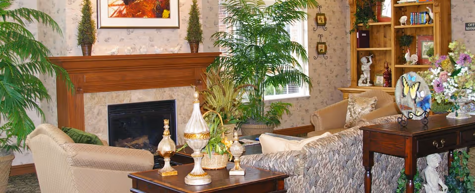 A cozy living room area with a wooden fireplace mantel decorated with two small potted plants and a framed picture above it. The room features comfortable upholstered sofas, a wooden side table with decorative vases, a large green potted plant near the window, and a wooden bookshelf filled with books and decorative items. A wooden console table holds a decorative plate with butterfly designs and a vase of flowers.