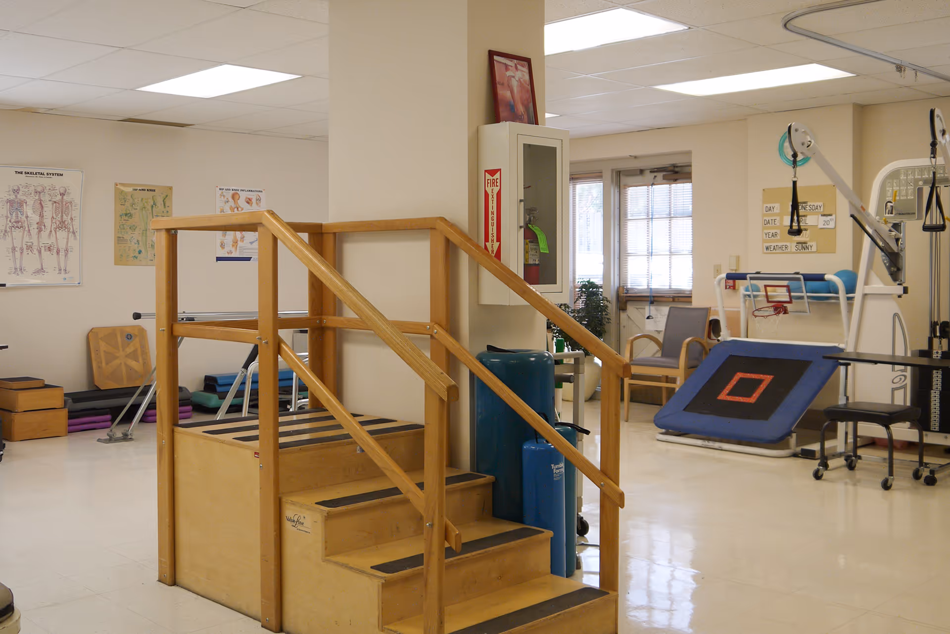 Physical therapy room with wooden stairs for rehabilitation, exercise equipment, anatomical posters on the wall, and chairs near a window with blinds.
