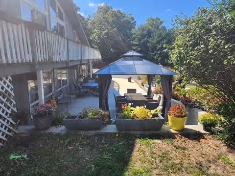 Outdoor patio area at Nichols Senior Care featuring a gazebo with a table and chairs underneath, surrounded by various potted plants and flowers. The patio is adjacent to a building with a balcony and windows, and trees are visible in the background under a clear blue sky.