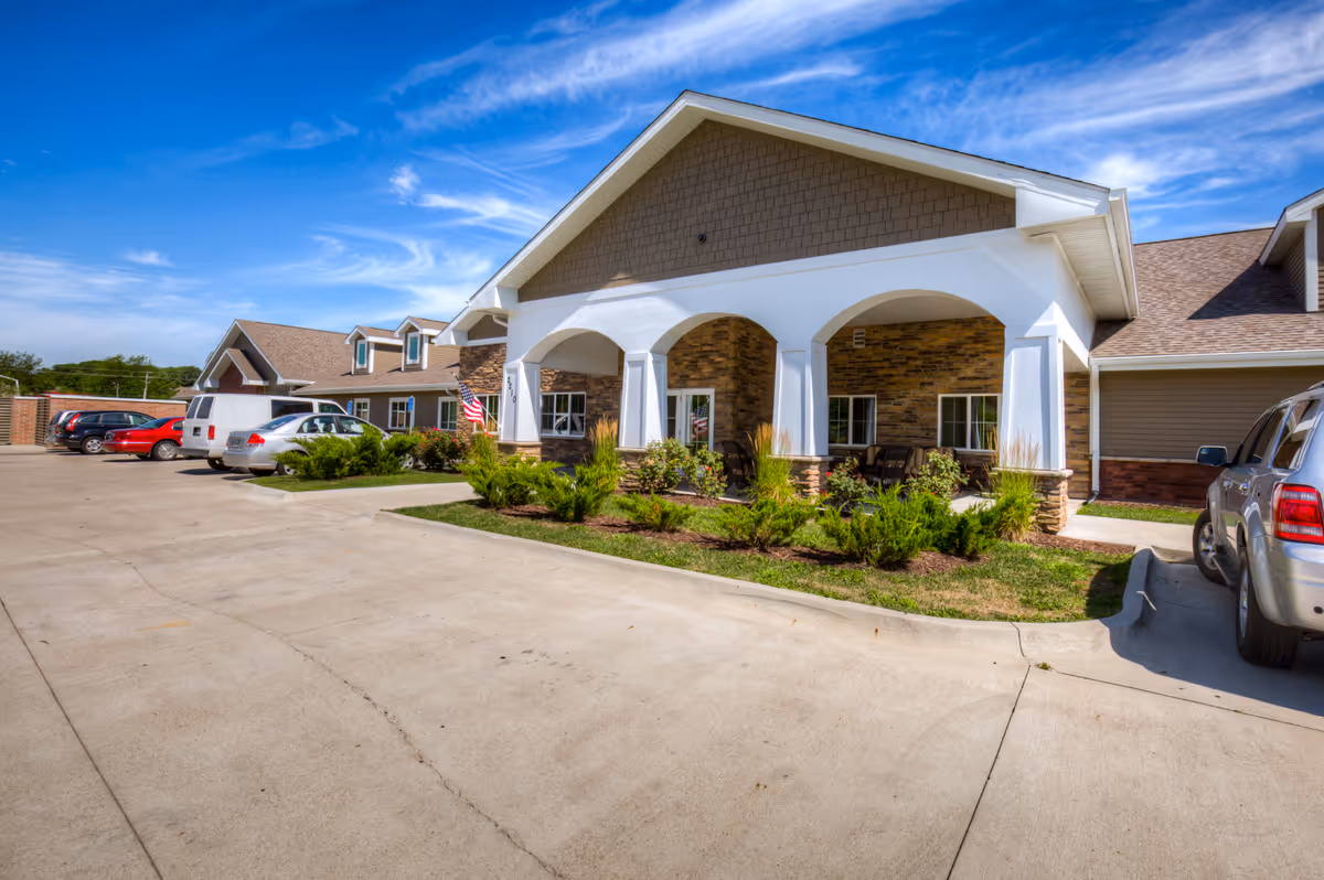 Front exterior of a single-story senior living facility with a covered arched entrance, landscaping, and parked cars.