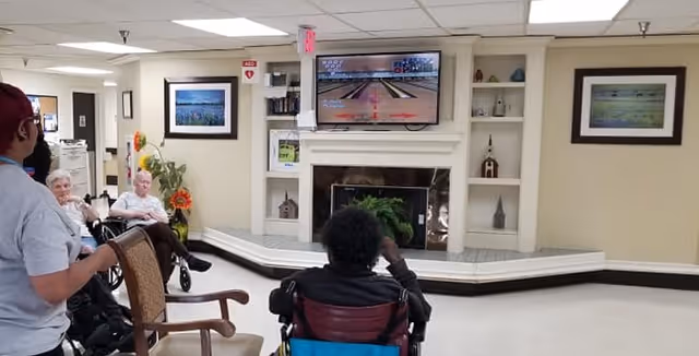 A group of elderly people, some in wheelchairs, are seated in a common area watching a television mounted above a fireplace. The room has beige walls with framed pictures and built-in shelves with decorative items. A staff member is standing nearby.