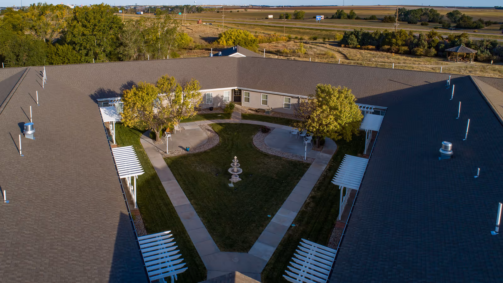 Aerial view of a single-story assisted living facility arranged in a U-shape around a central courtyard with a green lawn, a three-tiered fountain, paved walkways, and several trees providing shade. The surrounding area is rural with open fields and a gazebo visible in the distance.