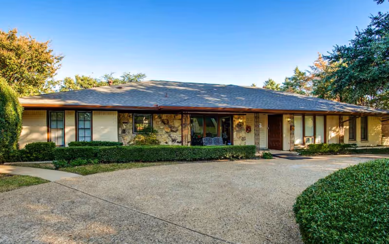 Single-story building with a stone and brick exterior, surrounded by neatly trimmed bushes and trees under a clear blue sky. A curved driveway leads to the entrance with a brown door and a small porch area with seating.