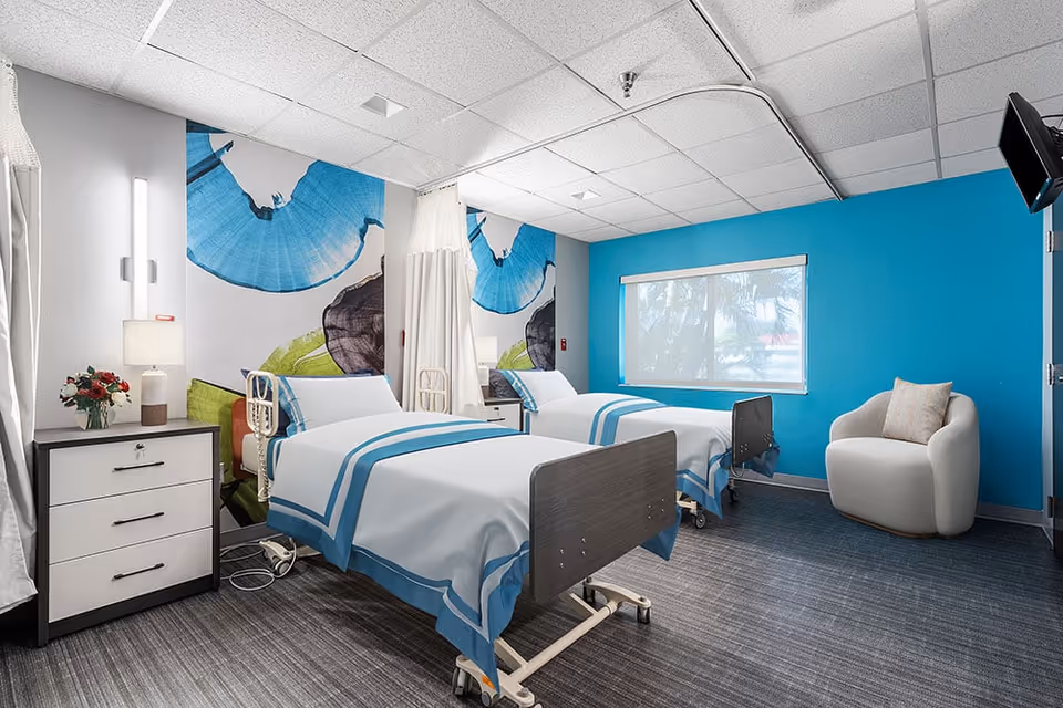 A bright and clean patient room in a nursing center featuring two hospital beds with white and blue bedding, a bedside table with a lamp and flowers, a comfortable armchair, a large window with a view outside, and colorful wall art with blue and green abstract designs.