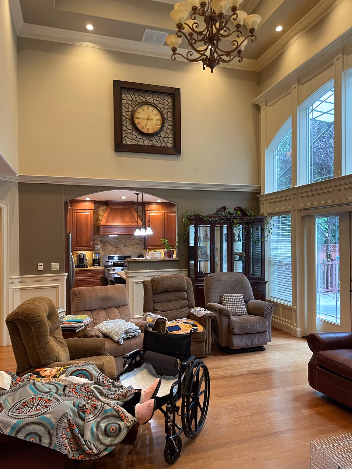 A cozy living room area with four recliner chairs arranged around a central space. One recliner has a colorful blanket draped over it and a wheelchair positioned nearby. The room features large windows with white blinds, a wooden floor, a chandelier hanging from a high ceiling, and a large decorative clock on the wall. In the background, there is a kitchen area with wooden cabinets and pendant lights, and a glass display cabinet with plants on top.