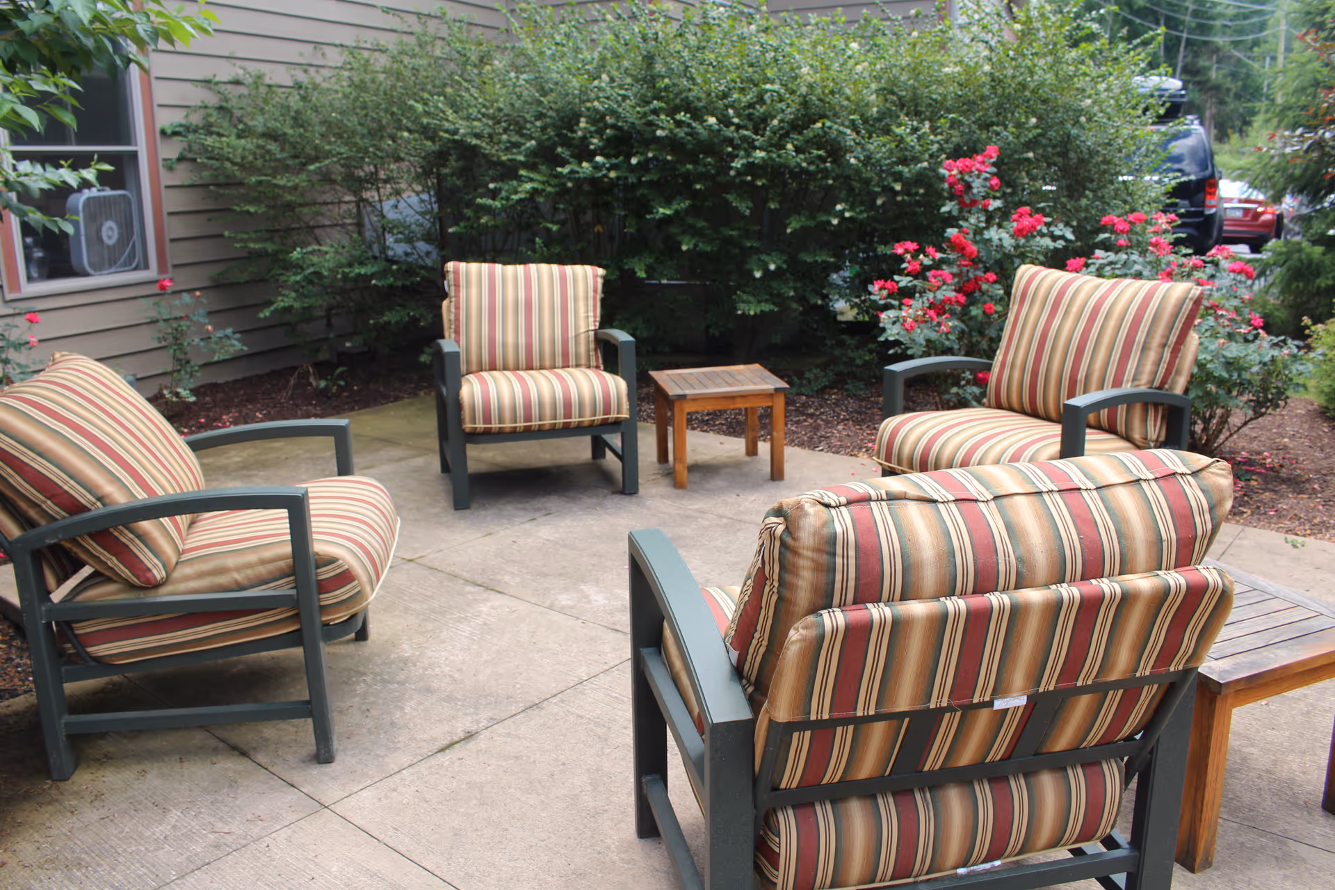 Outdoor patio seating area with four striped cushioned chairs and small wooden tables surrounded by shrubs and flowers.