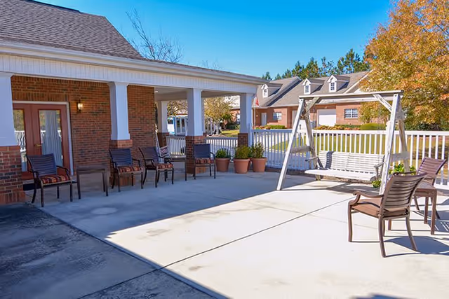 Outdoor patio area with several chairs arranged along a brick building with white columns. There is a white wooden swing and potted plants near a white fence. Trees with autumn foliage and additional buildings are visible in the background under a clear blue sky.
