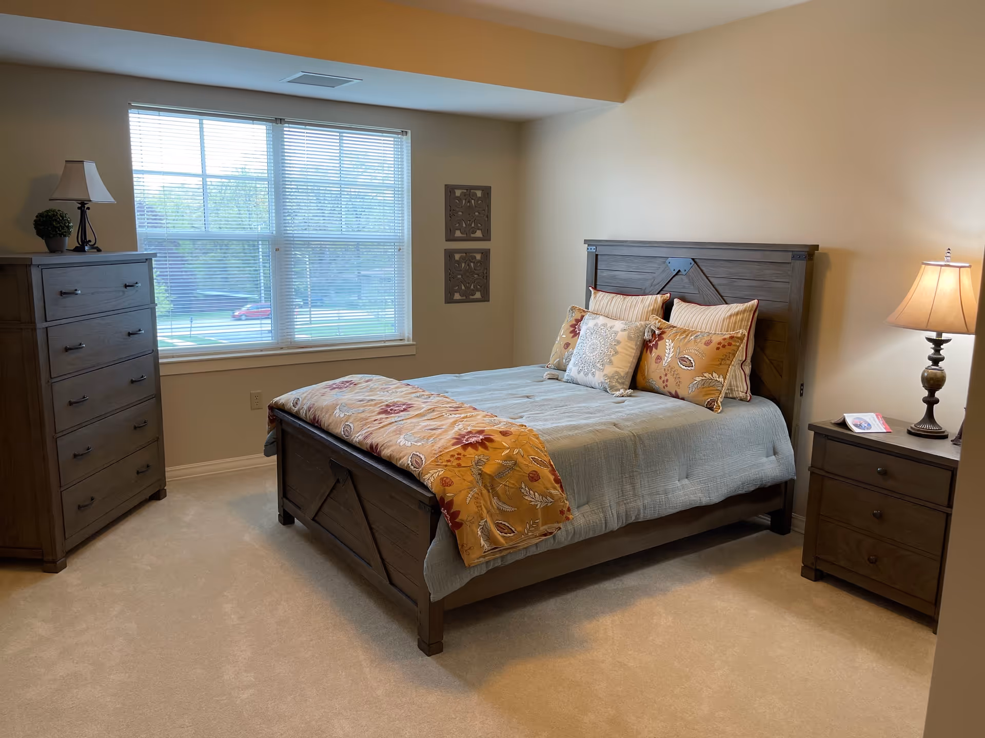A bedroom with a large window covered by blinds, a wooden bed with a headboard and footboard, decorated with multiple pillows and a quilt. There is a wooden dresser with a lamp and a small plant on top to the left of the window, and a wooden nightstand with a lamp and a brochure to the right of the bed. The walls are beige and the carpet is light-colored.