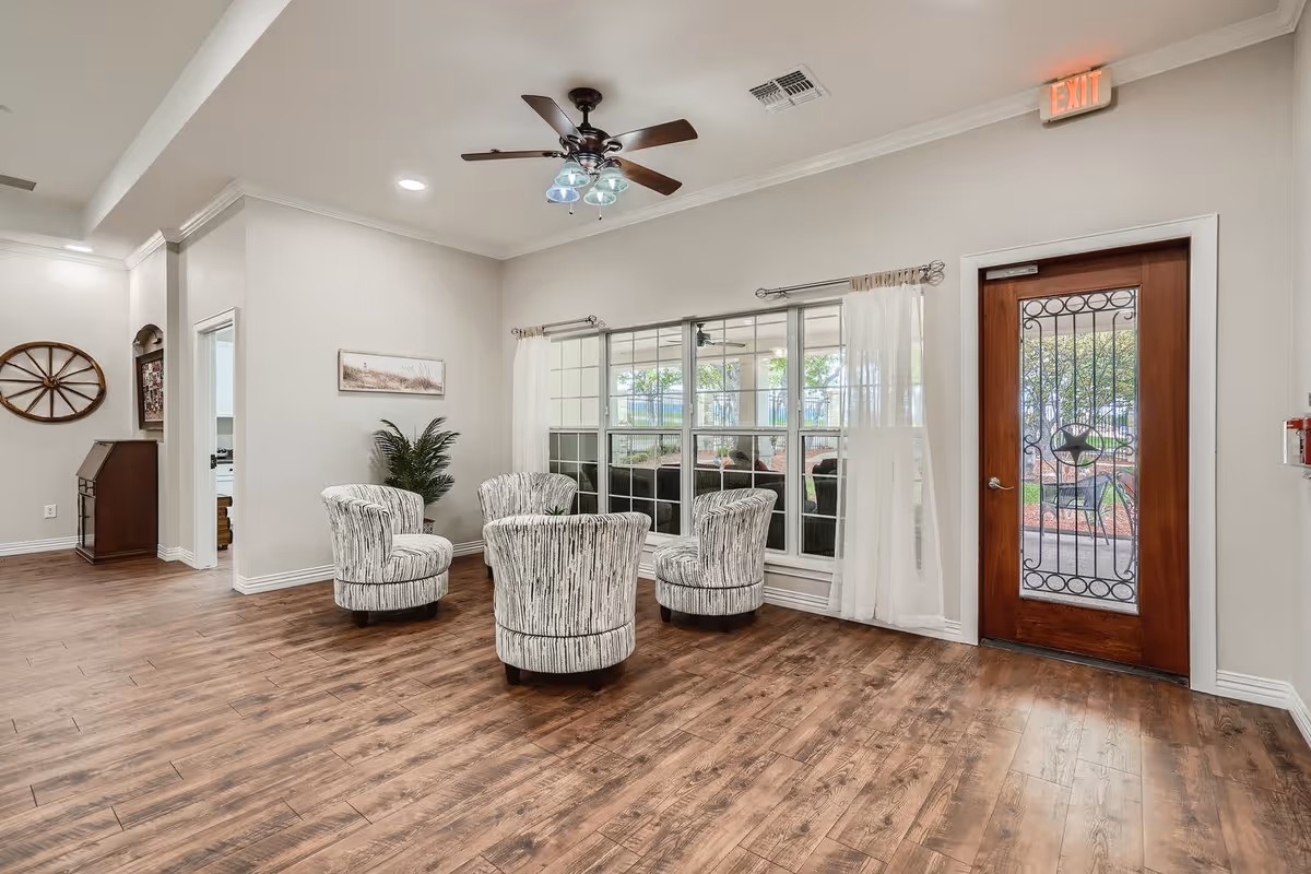 A bright and spacious senior living facility common area with four patterned armchairs arranged in a circle on a wooden floor. There is a large window with sheer white curtains letting in natural light, a wooden door with decorative glass, a ceiling fan with lights, and neutral-colored walls with minimal decor including a plant and a wall-mounted picture.
