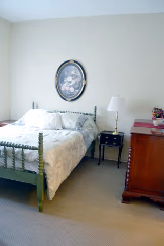 A bedroom with a green wooden bed frame, white and patterned bedding, a small black nightstand with a brass lamp, a wooden dresser with a floral arrangement, and a floral painting hanging on the light-colored wall.