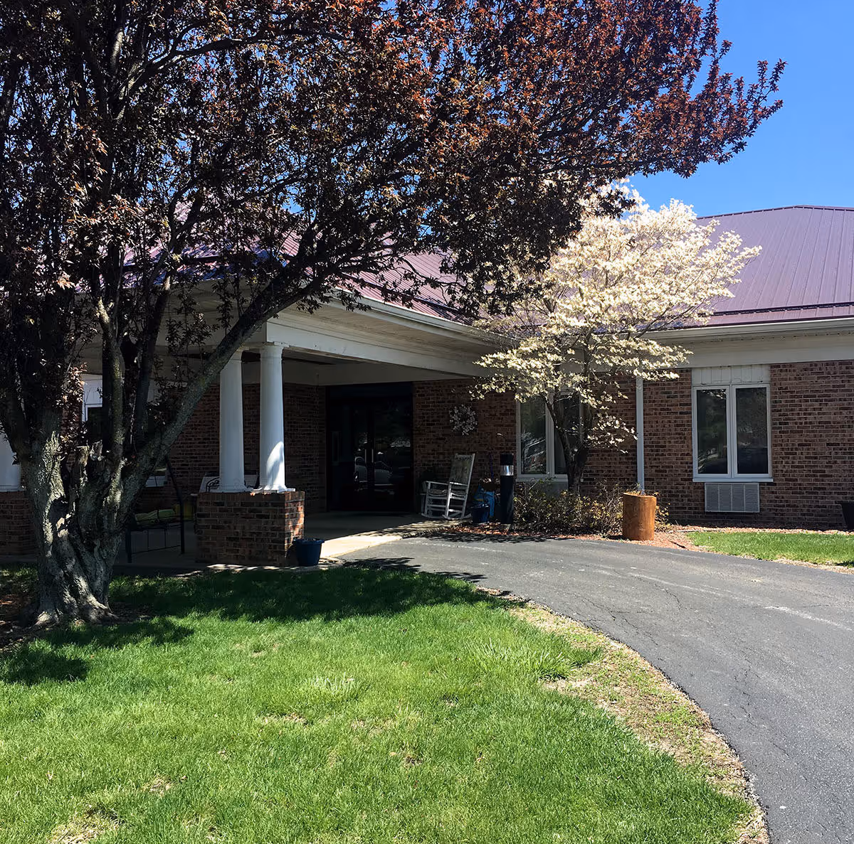 Front entrance of a brick nursing home with a covered portico, trees in bloom, and a curved driveway leading up to the door.