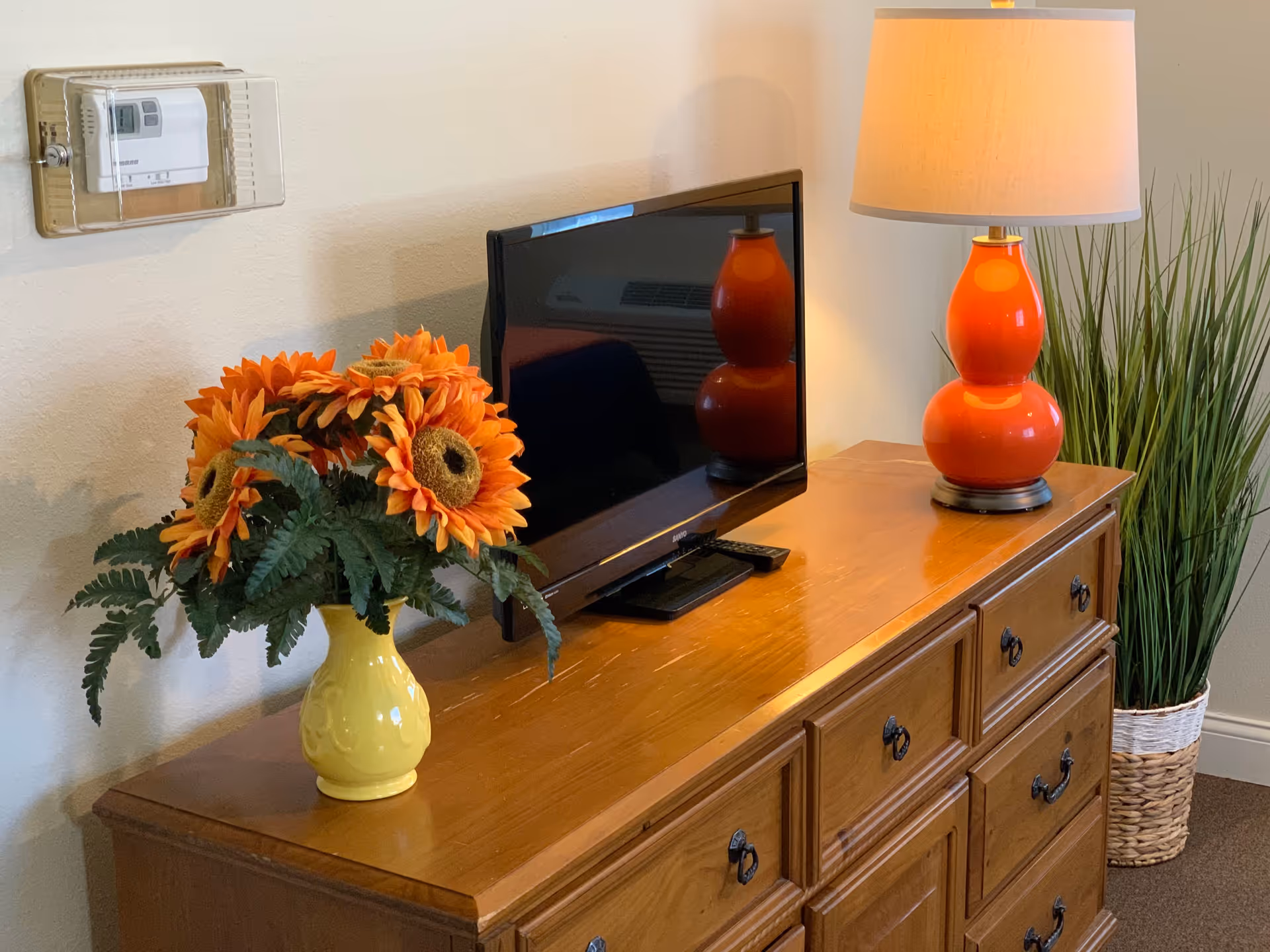 A wooden dresser with multiple drawers holds a flat-screen TV, a bright orange lamp with a beige lampshade, and a yellow vase filled with artificial sunflowers and green leaves. To the right of the dresser is a tall green plant in a woven basket. A thermostat is mounted on the wall above the dresser.