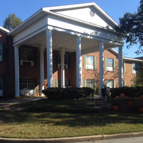 Front exterior view of a two-story brick building with white columns supporting a large covered entrance. There are bushes and a small lawn in front, along with some pumpkins and a sign that reads 'Immediate Move In'.