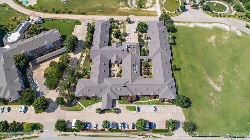 Aerial view of Morada Cedar Hill senior living facility showing multiple connected buildings with gray roofs, surrounded by green lawns, trees, parking lots with cars, and pathways.