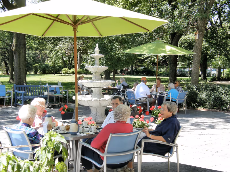 Several elderly people sitting at outdoor tables under green umbrellas, enjoying a sunny day in a garden-like patio area with a multi-tiered white fountain and surrounding trees and plants.