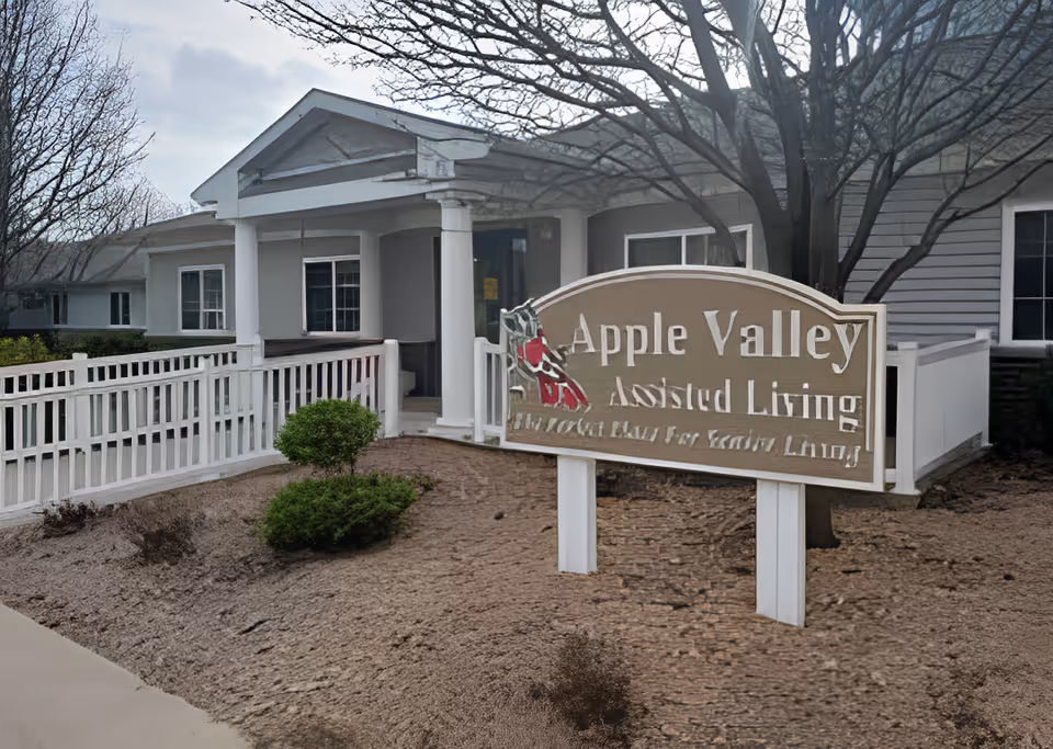 Exterior view of Apple Valley Assisted Living facility showing the building entrance with white columns, a white fence, some bushes, and a sign with the facility name in front.