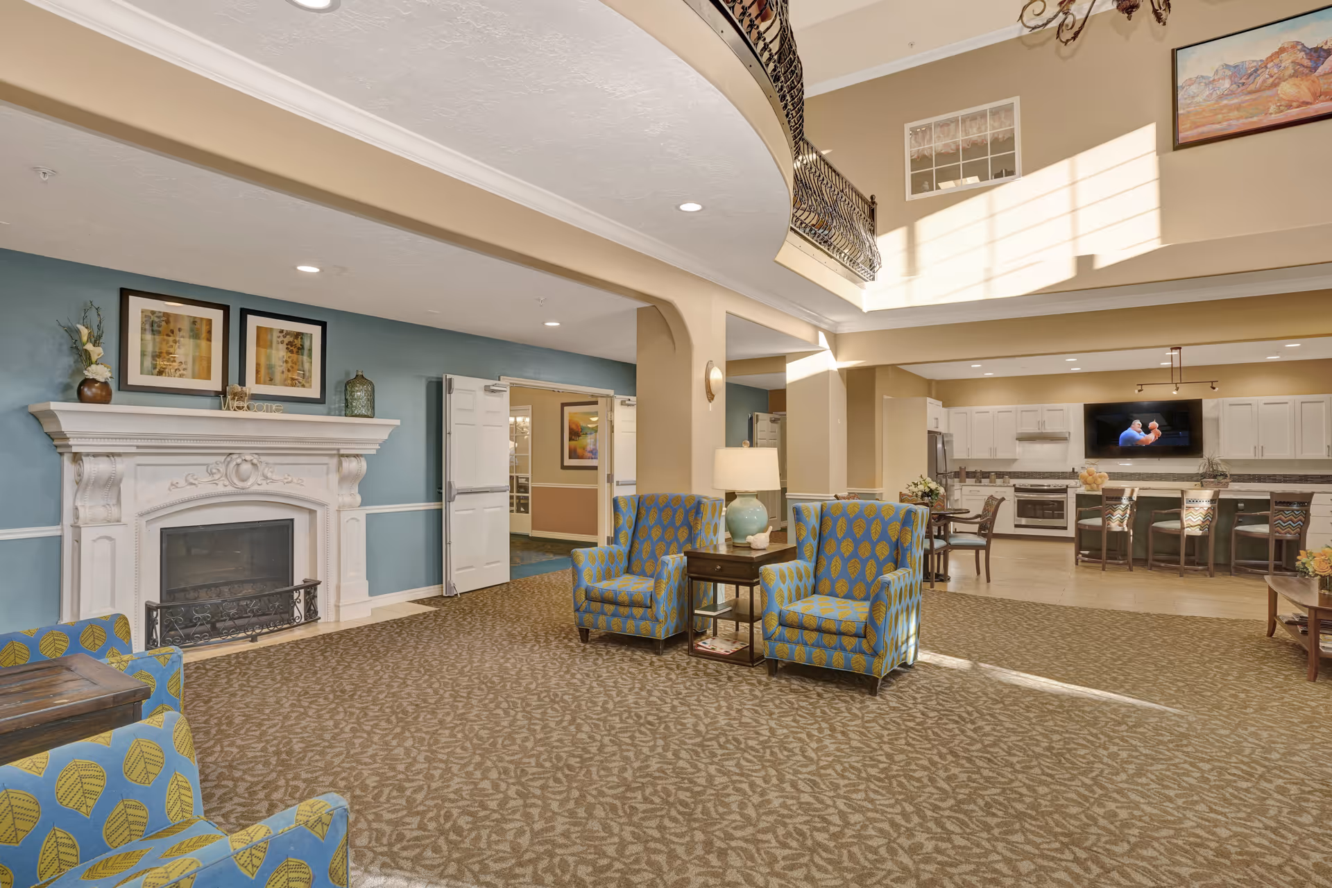 A spacious senior living facility common area with patterned carpet and beige walls. The room features a white decorative fireplace with framed artwork above it, several blue armchairs with yellow leaf patterns, a wooden side table with a lamp, and a kitchen area in the background with white cabinets, a stove, and a wall-mounted TV. Sunlight streams in through high windows, illuminating the space.