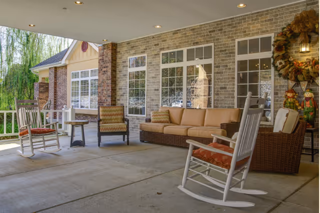 Covered outdoor patio area with cushioned wicker sofa, two white rocking chairs with orange cushions, an armchair, and small tables. The background shows a brick wall with windows and a decorative wreath with autumn-themed decorations.