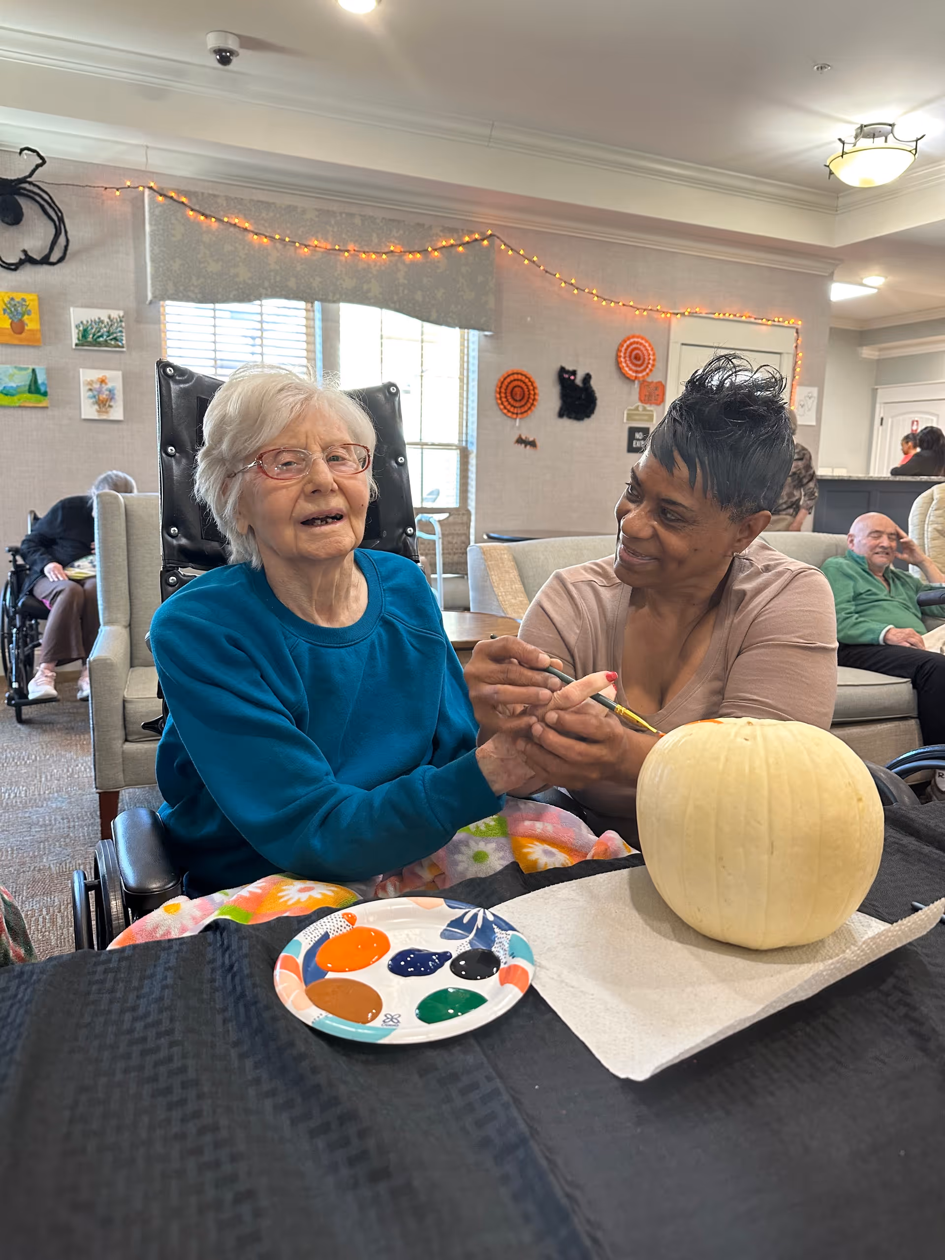 An elderly woman in a wheelchair is painting a white pumpkin with the assistance of a caregiver in a cozy common area decorated with Halloween-themed items and string lights. Other residents are visible in the background sitting on couches and chairs.