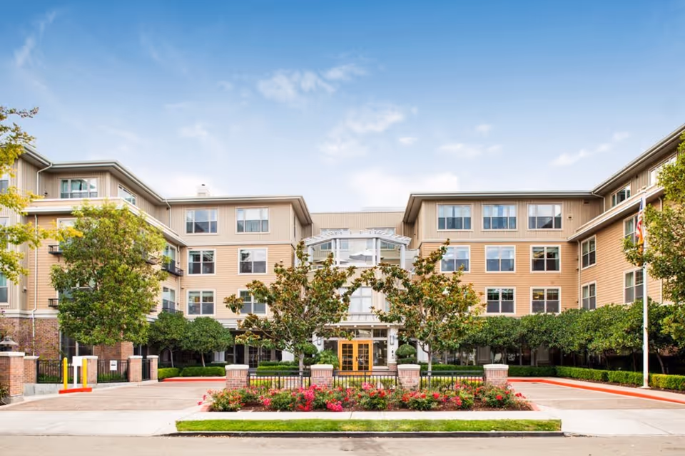 Front exterior view of a multi-story assisted living facility with beige siding and numerous windows, surrounded by well-maintained landscaping including trees, shrubs, and flowers under a partly cloudy sky.