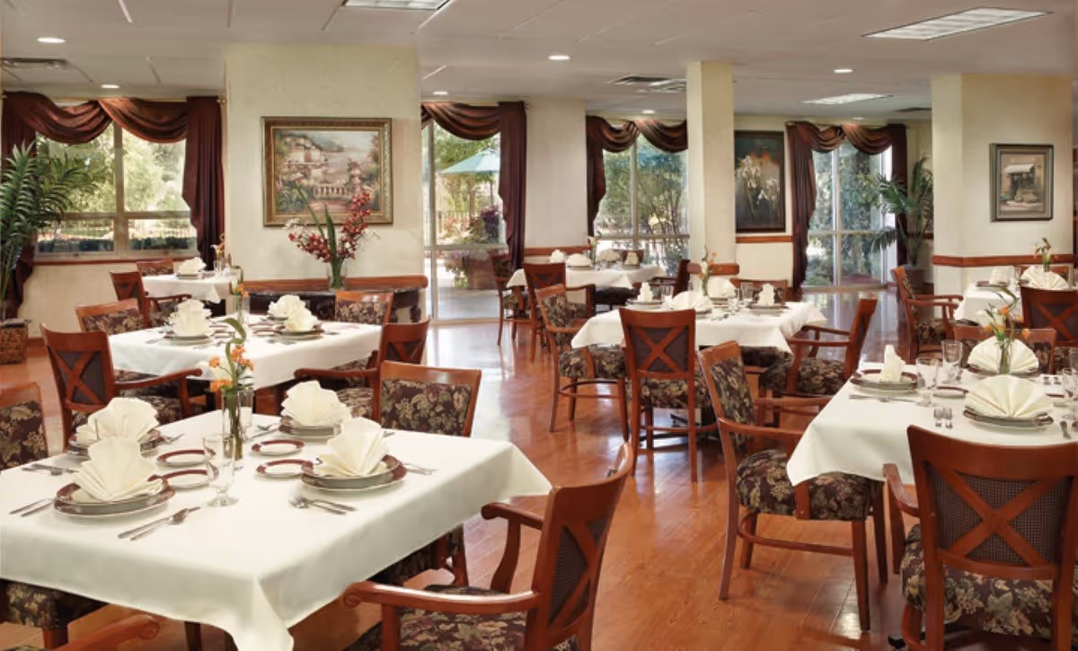 A well-lit dining room in Glenview Terrace with multiple tables covered in white tablecloths, each set with plates, silverware, glasses, and folded napkins. The chairs have wooden frames with floral-patterned cushions. Large windows with burgundy curtains allow natural light to fill the room, and there are framed paintings and plants decorating the space.