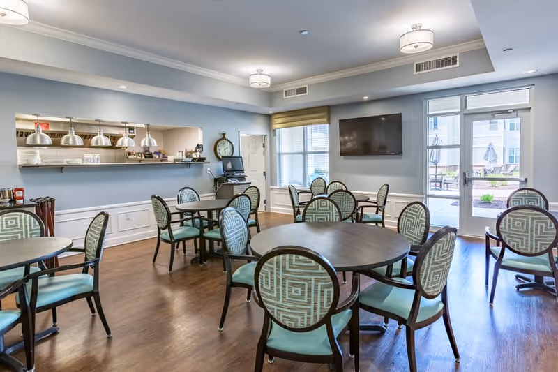 A bright dining room with several round tables and chairs featuring patterned backs and teal cushions. The room has light blue walls, wood flooring, and ceiling lights. A large window and glass door lead to an outdoor patio area with umbrellas and seating. A wall-mounted TV and a pass-through window to a kitchen area are also visible.